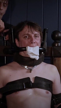 Ricky într-un fotoliu bondage, cu un căluș de oțel și umpluturi pentru șosete.